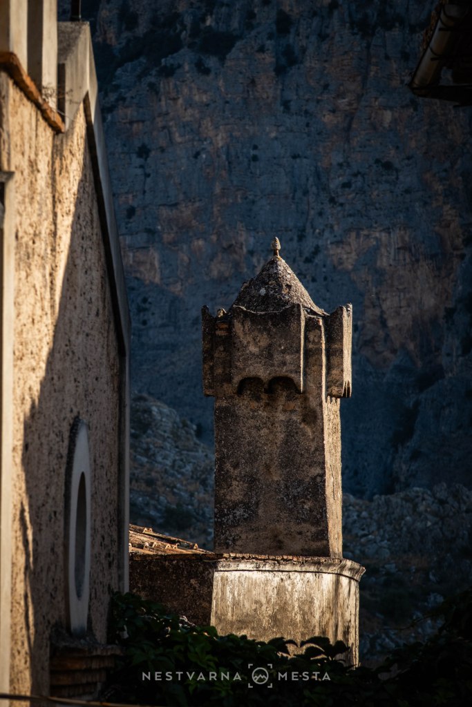 Close-up of a weathered stone chimney with a decorative top, set against a rocky mountainous backdrop.
