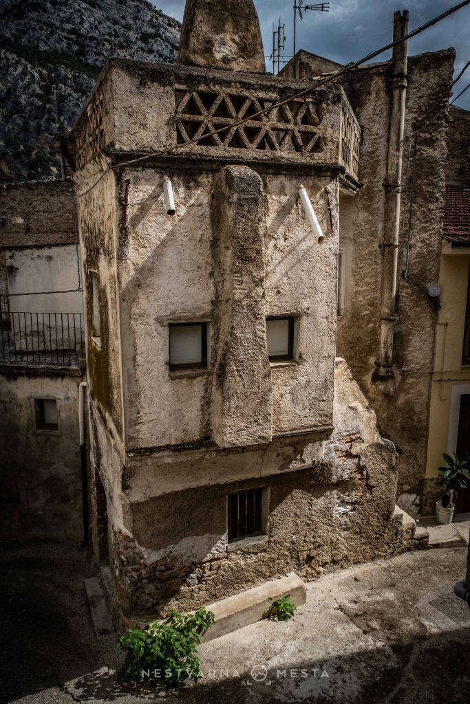 A weathered building with a unique architectural design resembling a face, featuring two windows for eyes and a protruding section that resembles a nose, set against a backdrop of rocky terrain under a cloudy sky.