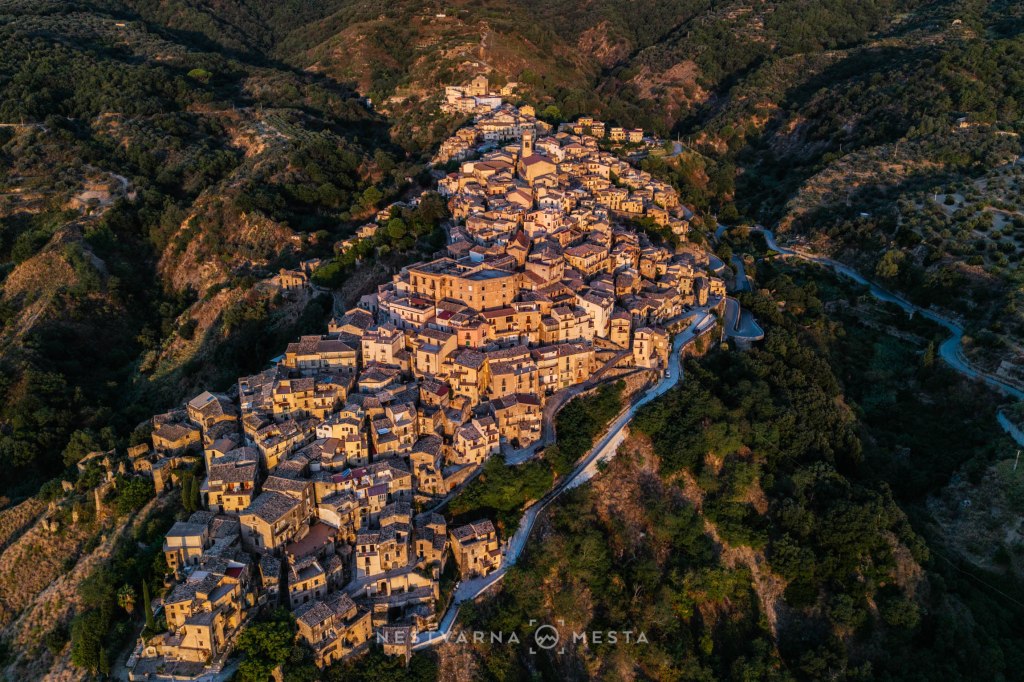 Aerial view of a picturesque hillside village with terracotta rooftops surrounded by lush greenery and rolling hills.