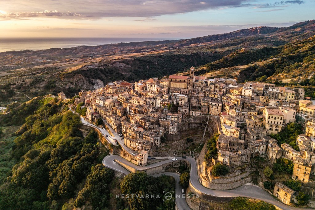 Aerial view of a hillside village with stone houses and narrow winding streets, overlooking a scenic coastline and green hills at sunset.