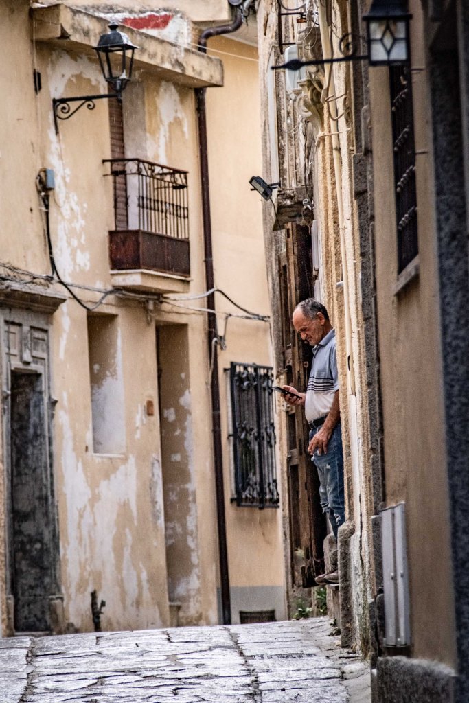 A man standing in an alleyway, looking at his phone, with weathered buildings and lanterns in the background.