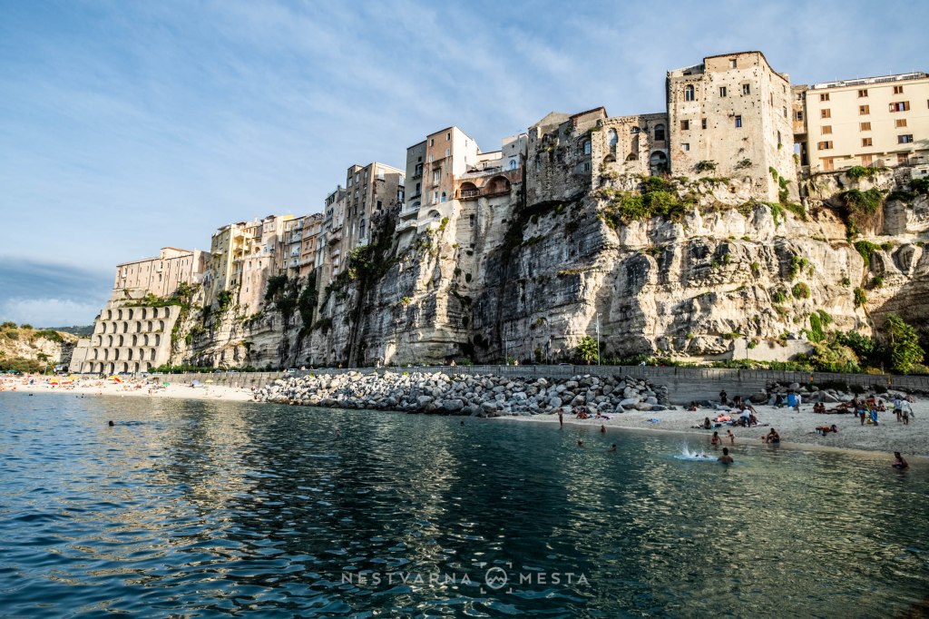 A scenic view of a rocky cliffside with buildings perched on top, overlooking a sandy beach and clear waters. People are swimming and relaxing along the shore.