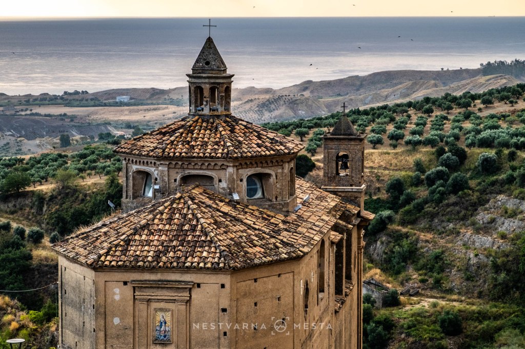 A scenic view of an ancient church with a tiled roof, surrounded by rolling hills and olive groves, overlooking the sea.