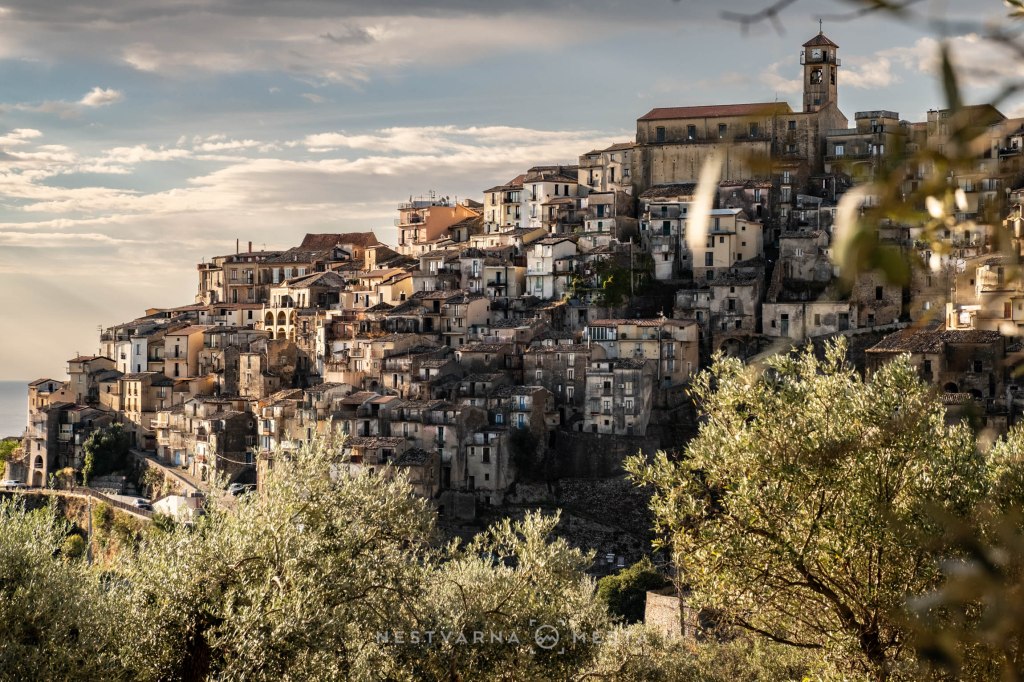 A scenic view of a hillside town with stacked buildings, olive trees in the foreground, and a cloudy sky in the background.