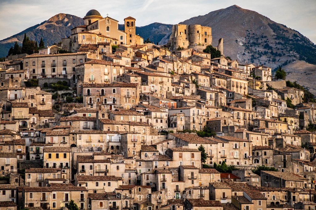 A picturesque hilltop village with clustered historic buildings, set against a backdrop of mountainous terrain.