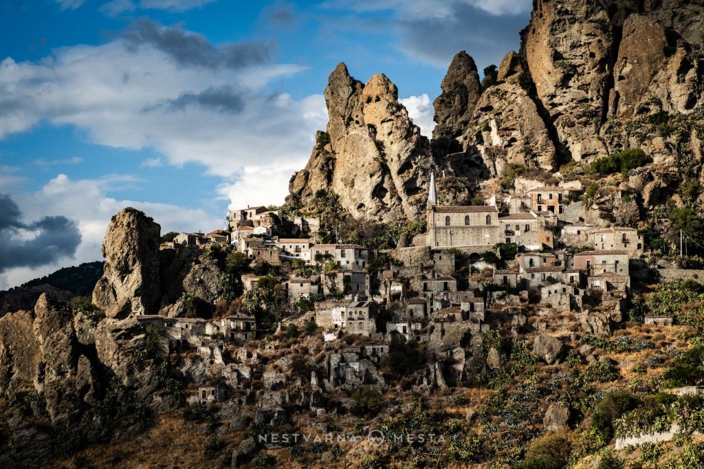 A panoramic view of a historic village built on rocky terrain, surrounded by dramatic cliffs and blue skies with scattered clouds.