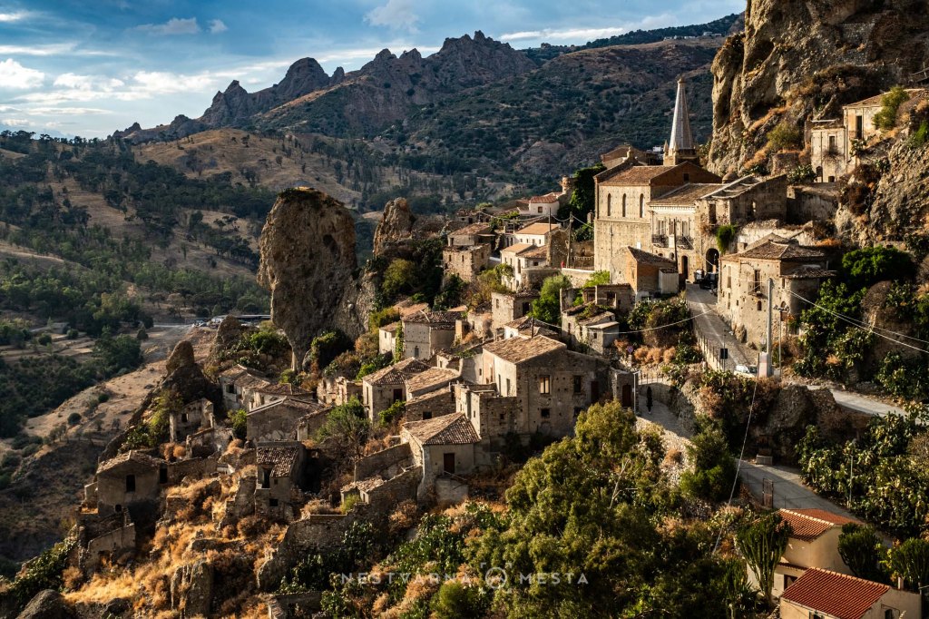 A scenic view of a historic village built into rocky terrain, featuring stone houses and lush greenery, set against mountains in the background under a partly cloudy sky.