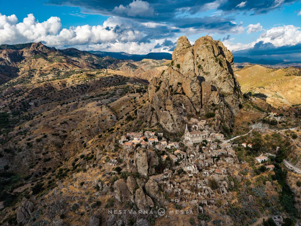 Aerial view of a small village nestled among rocky cliffs and mountains, featuring abandoned and historical buildings under a partly cloudy sky.