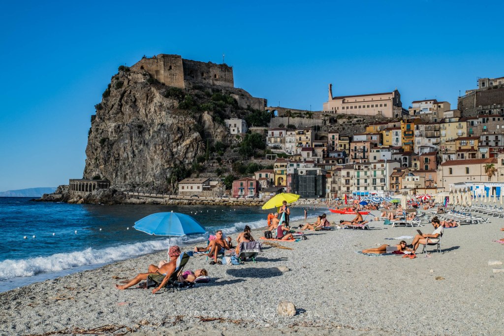 A beach scene featuring people lounging on the pebbled shore, colorful umbrellas, and rocky cliffs with a historic castle and buildings in the background under a clear blue sky.