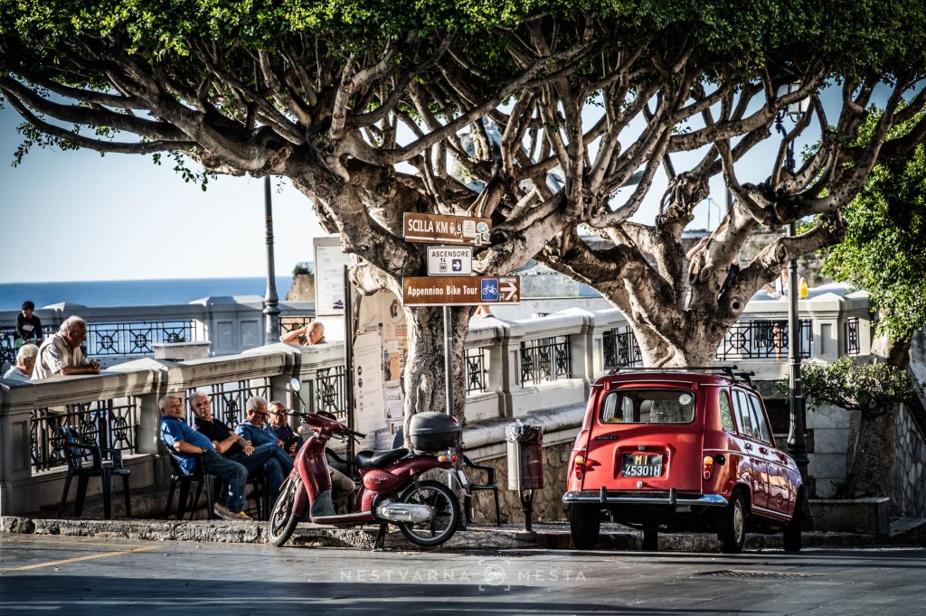 A vibrant red vintage car parked beside a scooter on a sunny street lined with stone walls and large trees. In the background, people are sitting on benches, enjoying the view of the sea, while directional signs indicate nearby destinations.