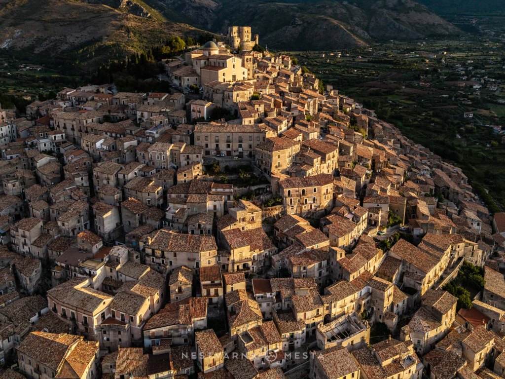Aerial view of a historic hilltop village with stone houses and terracotta roofs, surrounded by mountains and green valleys.