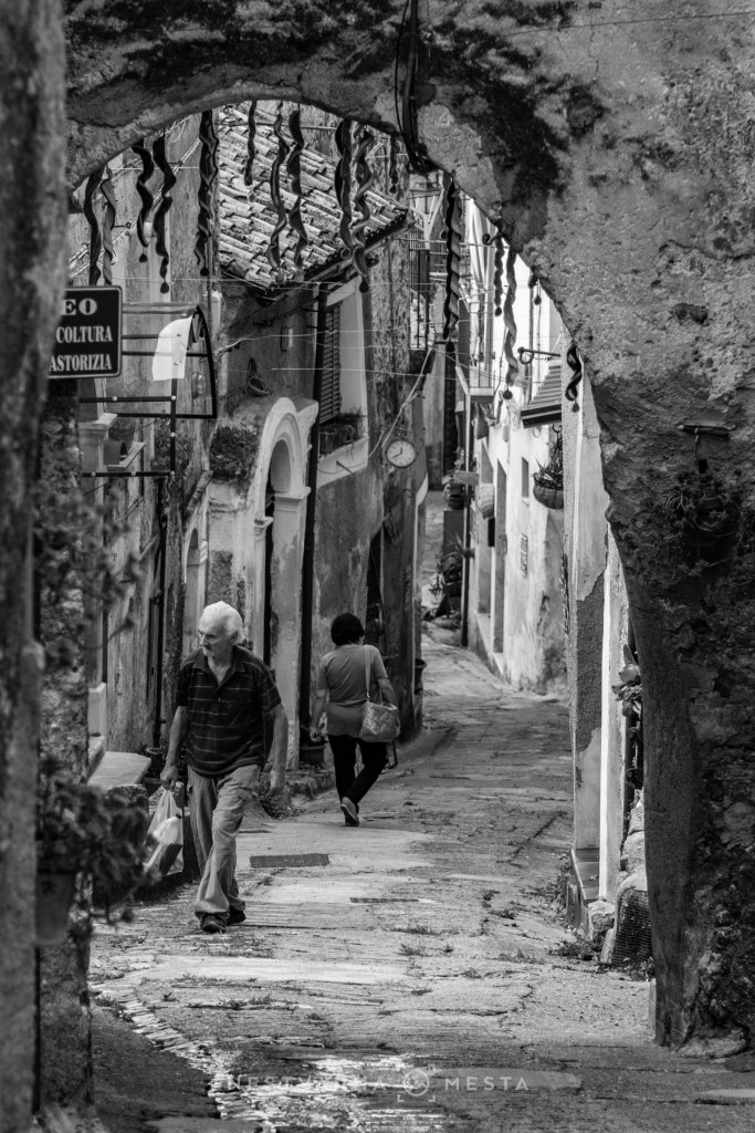 A narrow cobblestone street in a village, featuring an archway overhead. An elderly man walks with shopping bags, while a woman walks in the opposite direction. The scene is in black and white.