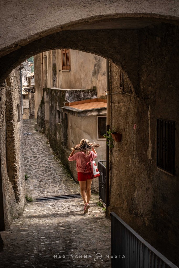 A woman with long hair walking down a cobblestone street in an old town, wearing a pink top and red shorts, with a striped bag slung over her shoulder.