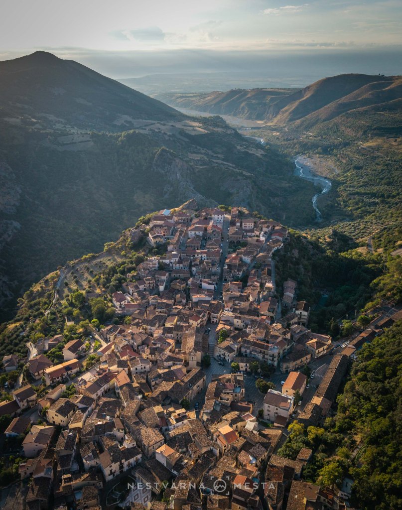 Aerial view of a historic village nestled in a mountainous landscape, surrounded by greenery and hills, with a winding river flowing nearby.