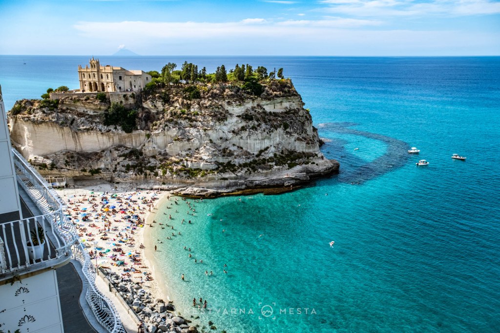 A scenic view of a beach with colorful umbrellas and people sunbathing, situated near a rocky cliff topped with a historic building, against a backdrop of clear blue water and a distant island.