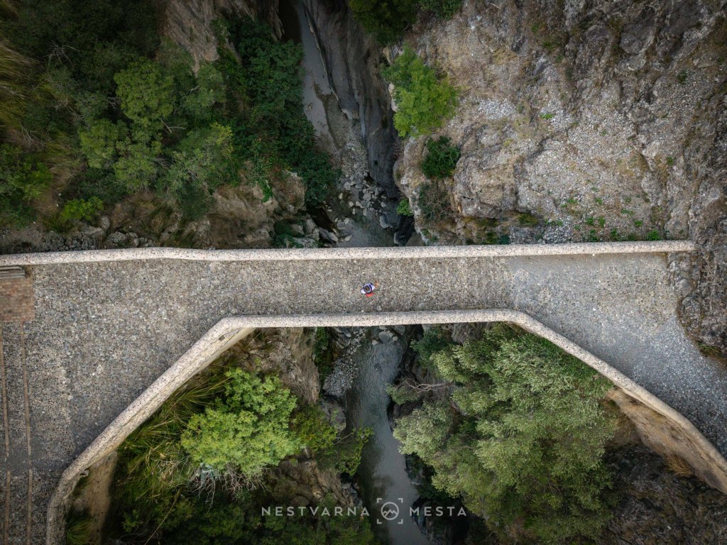 Aerial view of a stone bridge spanning a narrow canyon, surrounded by lush greenery and rocky terrain.