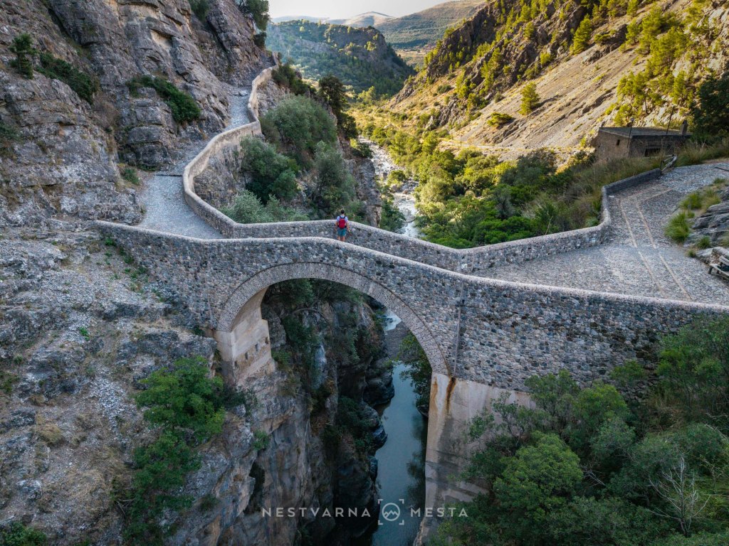 Aerial view of a stone arch bridge over a river, surrounded by rocky cliffs and lush greenery in a mountainous landscape.