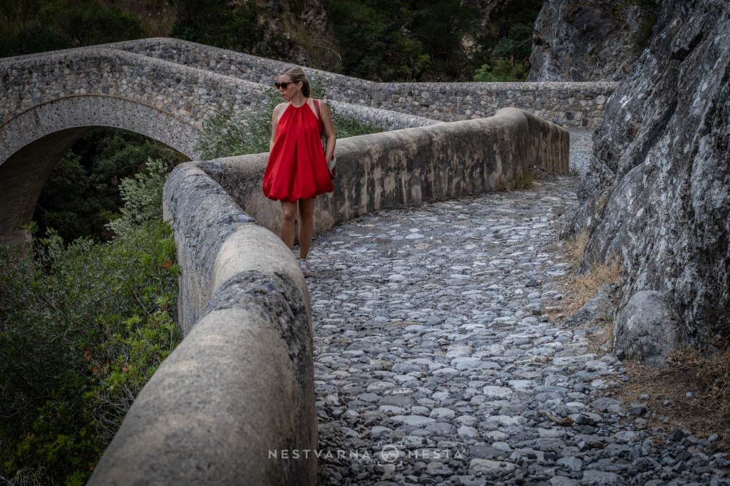 A woman in a red dress walking along a stone pathway with a curved stone railing, surrounded by greenery and a bridge in the background.