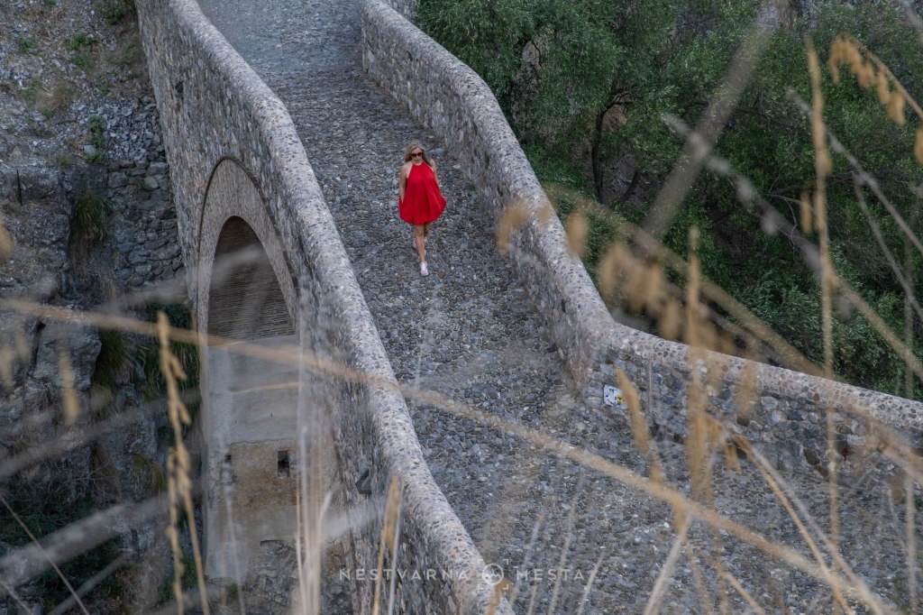 A woman in a red dress walking on a stone bridge surrounded by greenery.