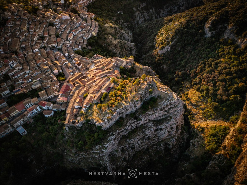 Aerial view of a mountainous landscape featuring a town with a series of terracotta-roofed buildings nestled among lush greenery.