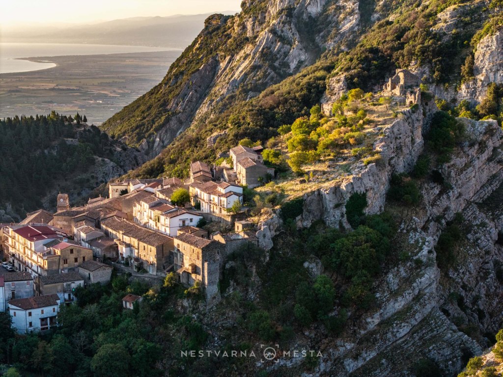 Aerial view of a picturesque hillside village nestled against a rocky cliff, with lush greenery and a distant view of the sea.