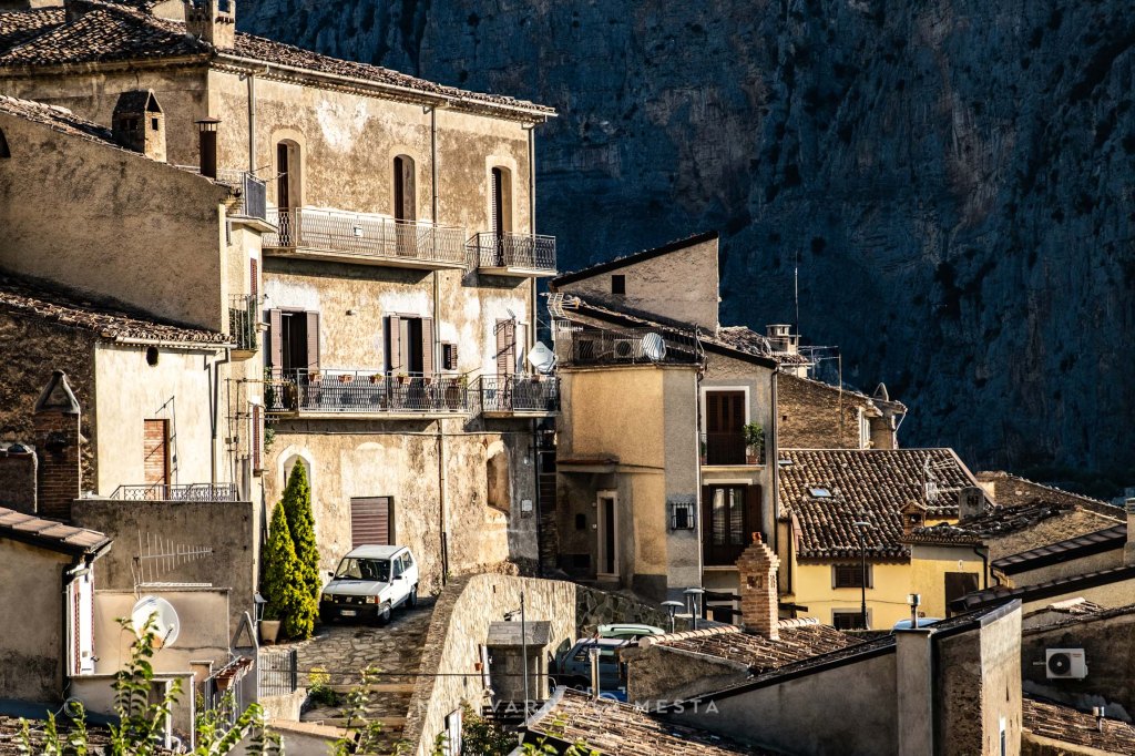 A scenic view of an Italian village with stone houses, balconies, and a parked white car, set against a rugged mountainous backdrop.
